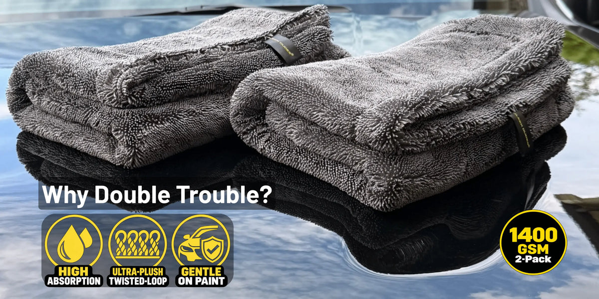 Two folded gray plush microfiber towels displayed on a reflective dark surface with water in the background. Text overlays read 'Why Double Trouble?' and '1400 GSM 2-Pack,' with feature icons for high absorption, ultra-plush twisted-loop texture, and gentle on paint.