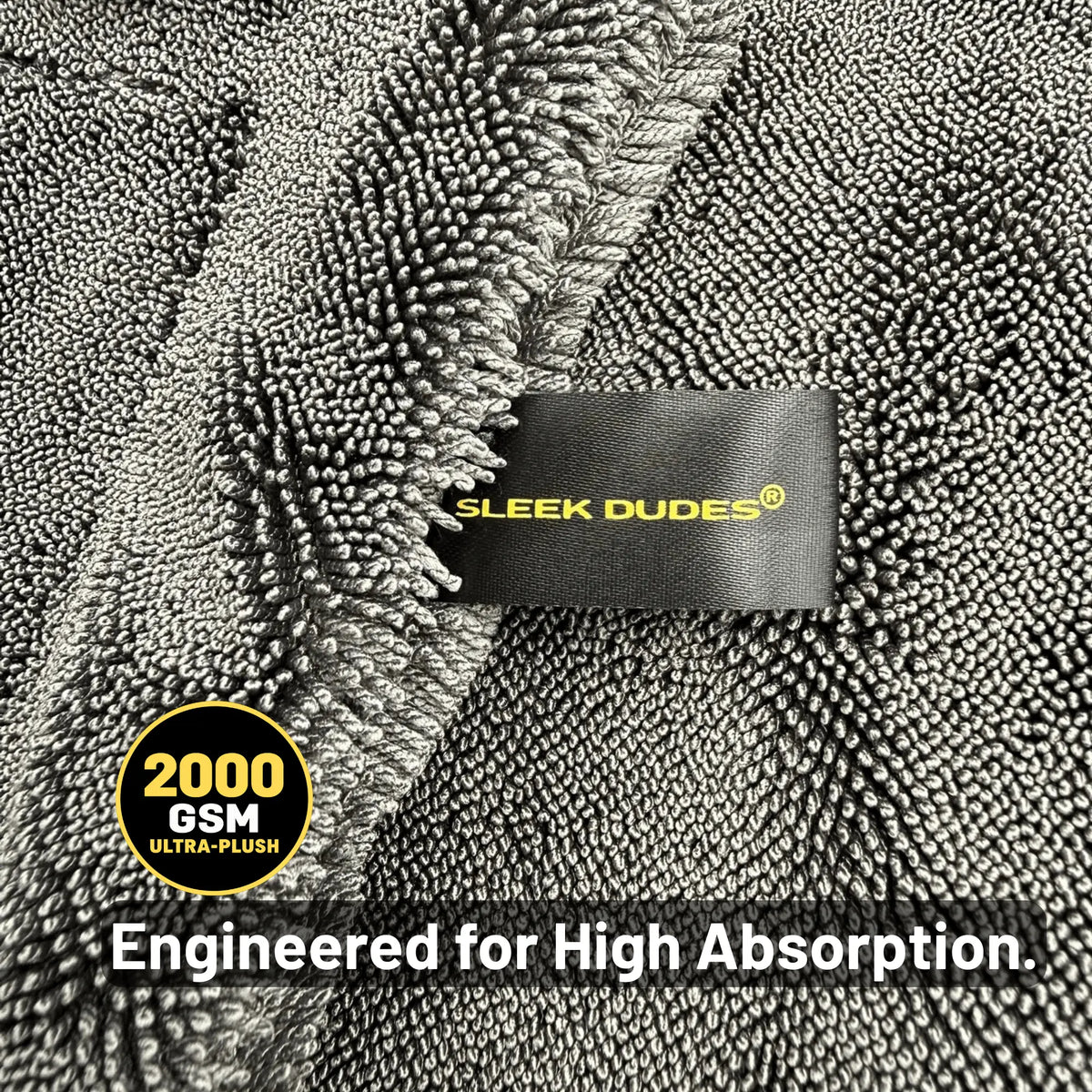 Close-up of a gray plush car drying microfiber with a black fabric tag reading 'SLEEK DUDES' in yellow. A circular badge says '2000 GSM ULTRA-PLUSH' and text at the bottom reads 'Engineered for High Absorption.