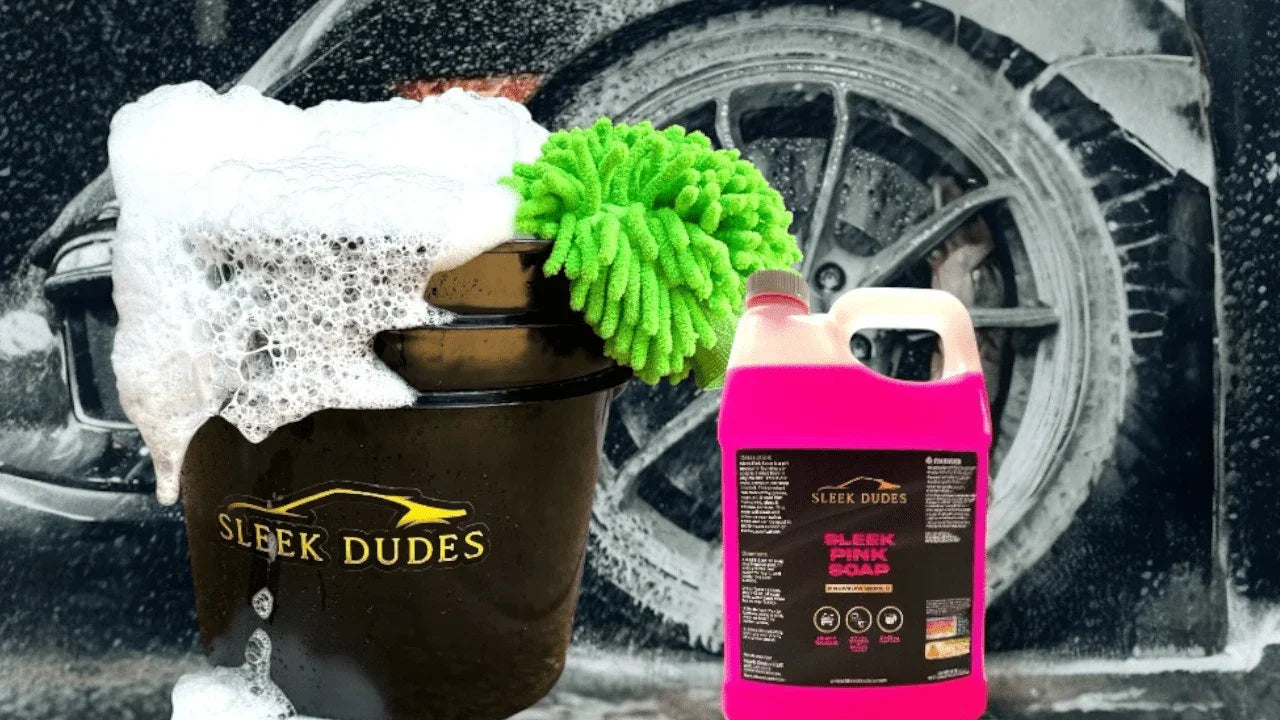 Sleek Dudes car wash kit featuring a foam-filled wash bucket, bright green chenille microfiber mitt, and Sleek Pink Soap gallon jug, with a foam-covered black car wheel in the background.