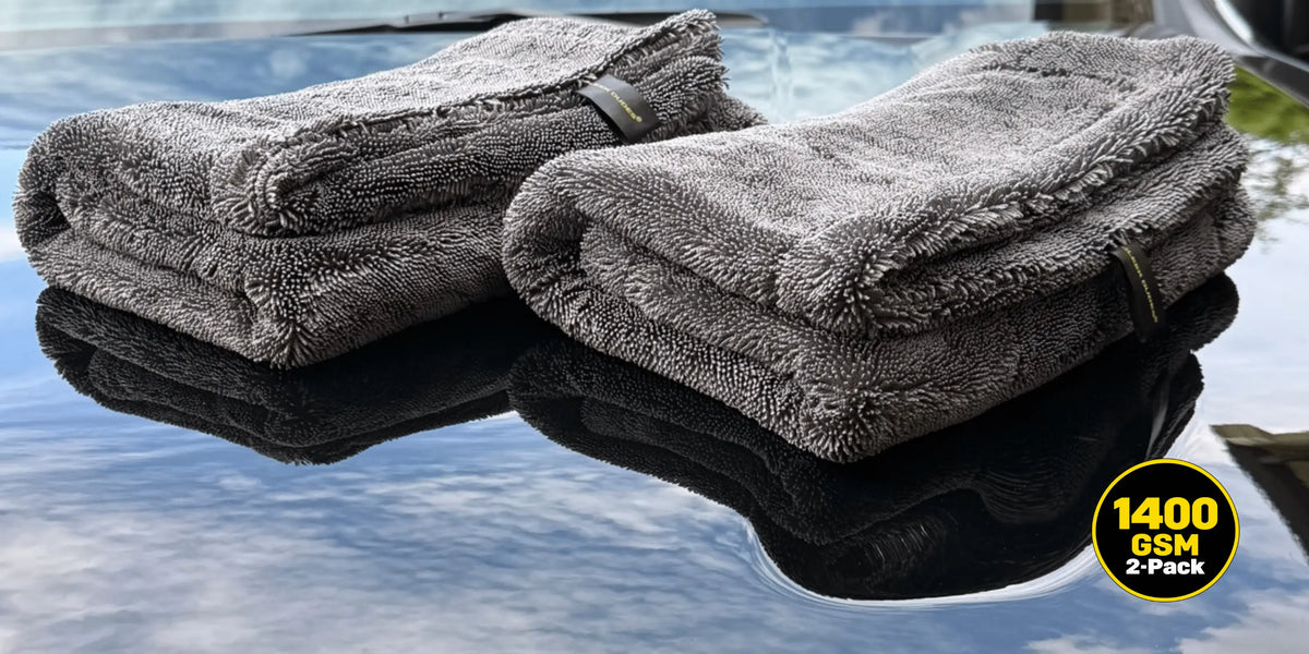 Two folded gray microfiber car drying towels resting on a reflective dark surface, with a yellow circular badge in the bottom right reading 1400 GSM 2-PACK.