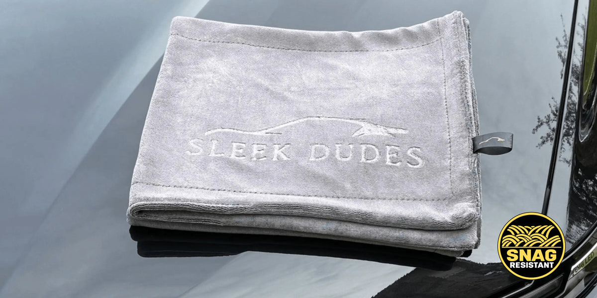 A folded light gray microfiber car towel with 'SLEEK DUDES' embroidered on the front, resting on a glossy dark car hood. A circular 'SNAG RESISTANT' badge is visible in the bottom right corner.