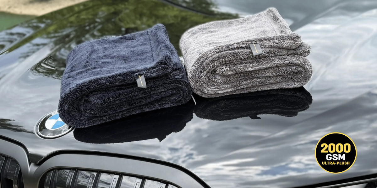 Two folded microfiber drying towels, one black and one light gray, displayed on the glossy black hood of a BMW car. A gold circular badge in the bottom right corner reads '2000 GSM ULTRA-FINE'.