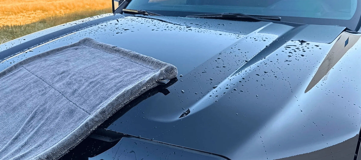 A gray microfiber towel draped over the front hood of a dark car outdoors, with water droplets visible on the glossy hood surface.
