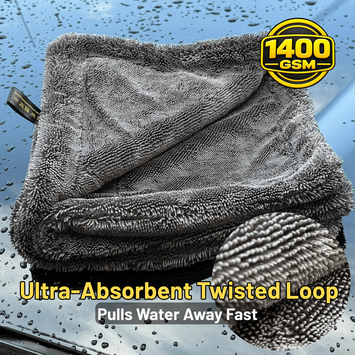 A folded dark gray microfiber cleaning cloth with a twisted loop texture, shown on a wet blue surface with raindrops. A circular badge reads 1400 GSM, and text at the bottom says Ultra-Absorbent Twisted Loop and Pulls Water Away Fast.