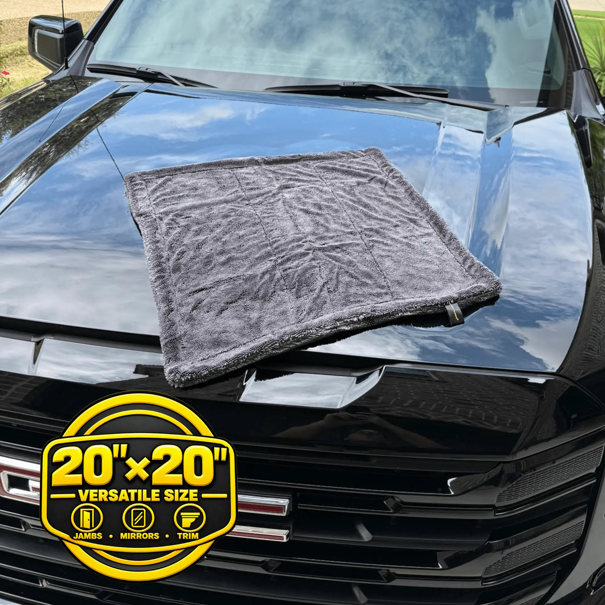 A dark gray microfiber cleaning cloth laid on the hood of a black vehicle, with a yellow circular badge at the bottom left reading 20x20 Versatile Size.