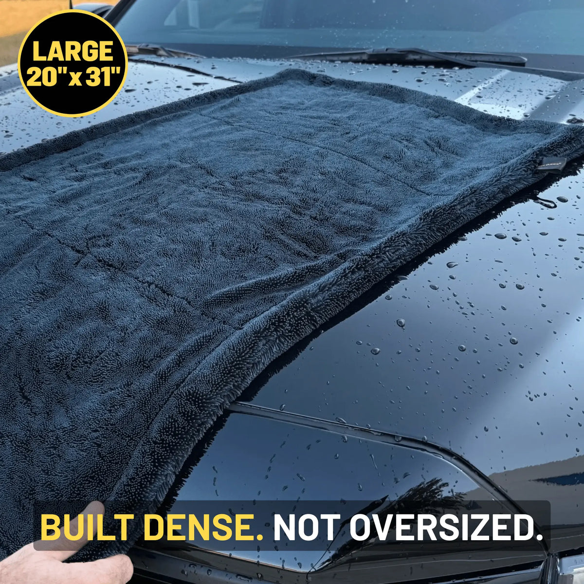 A large dark gray microfiber car drying towel laid across the hood of a wet car, with a hand holding the corner to show flexibility. Text overlays read 'LARGE 20" x 31"' and 'BUILT DENSE.