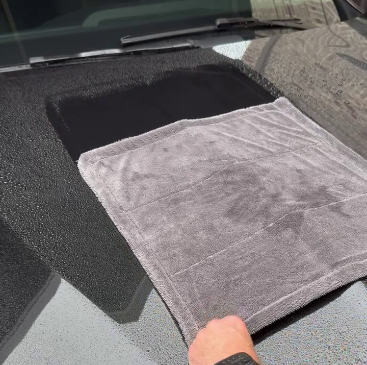 A gray microfiber towel is being held over a black car hood, with a person's hand visible at the bottom.